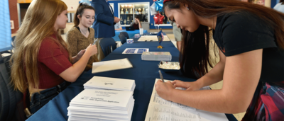 Two students, seated at a table covered in documents, facilitating a registration drive while another student fills out a sheet of paper across from them.